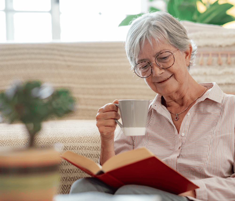 Elderly woman with glasses, holds a cup and reads a book, sitting on the floor in a cozy living room. - Home Instead