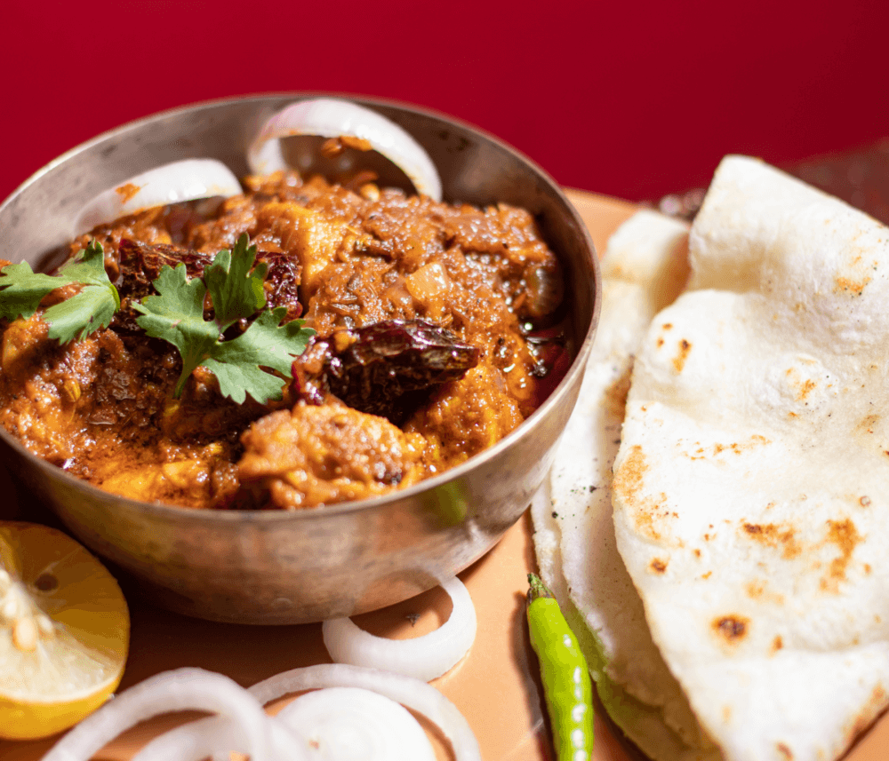 A bowl of spicy curry garnished with cilantro, served with naan bread, sliced onions, chili peppers, and a lemon wedge. - Home Instead
