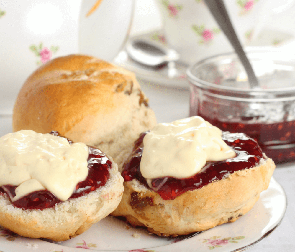 Bun with jam and cream halves on plate, whole bun, jar of jam, and floral teacup in background. - Home Instead