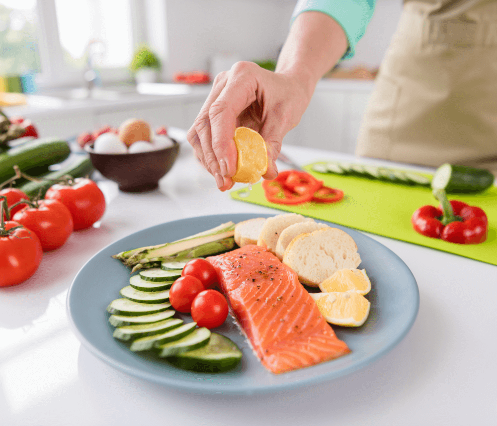 Person squeezing lemon over a plate with salmon, sliced cucumbers, tomatoes, asparagus, and bread in a kitchen. - Home Instead