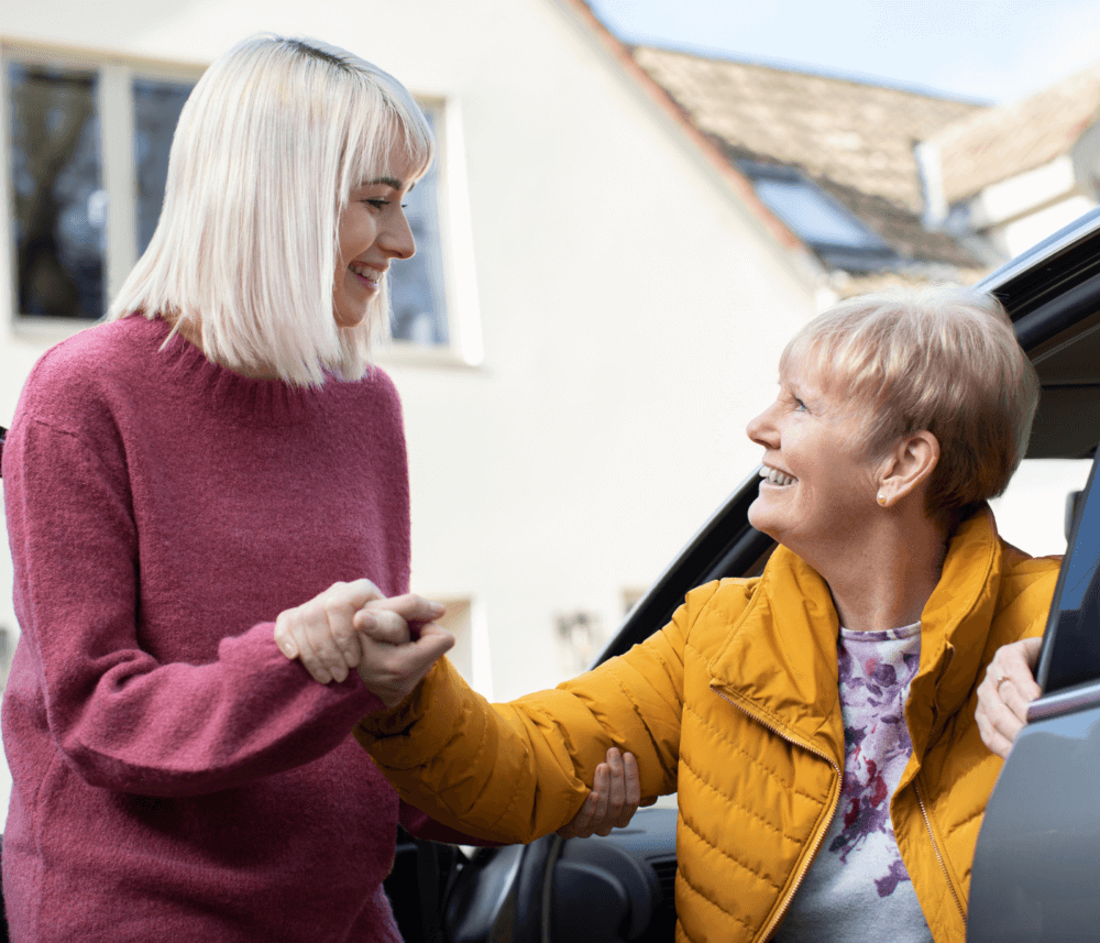 A younger person helps an older woman out of a car, both smiling, with a house in the background. - Home Instead