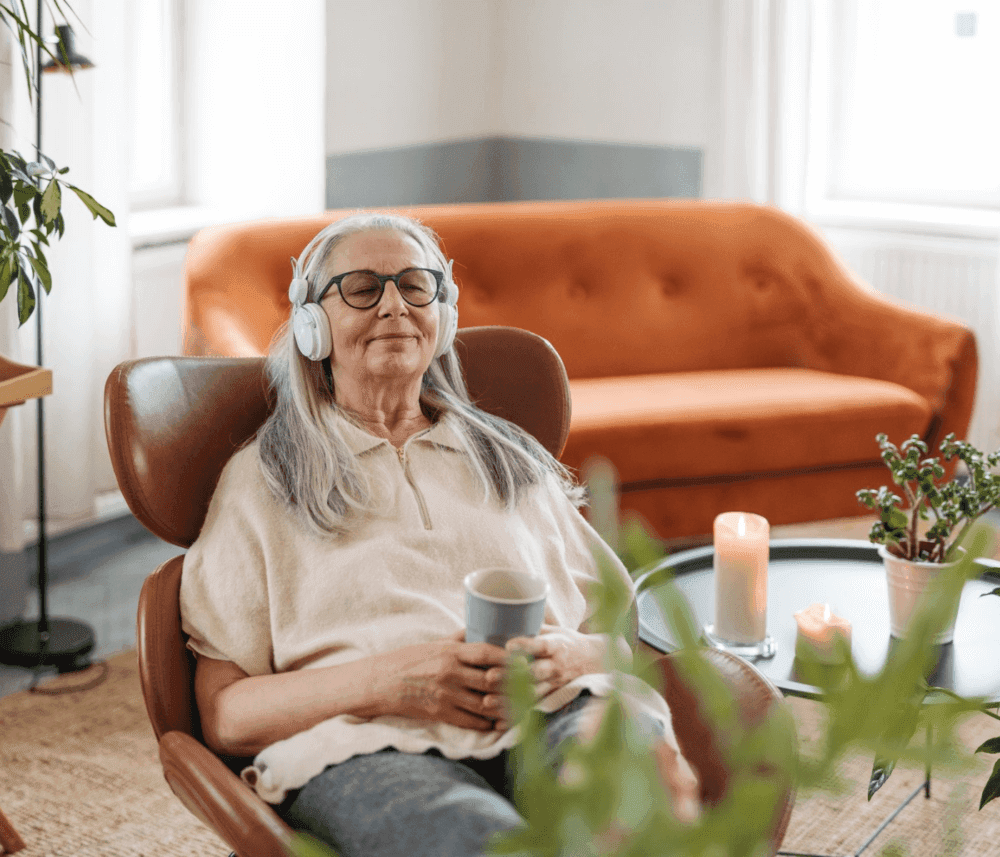 An elderly woman with headphones, holding a cup, relaxes in a cozy living room with an orange sofa and plants. - Home Instead
