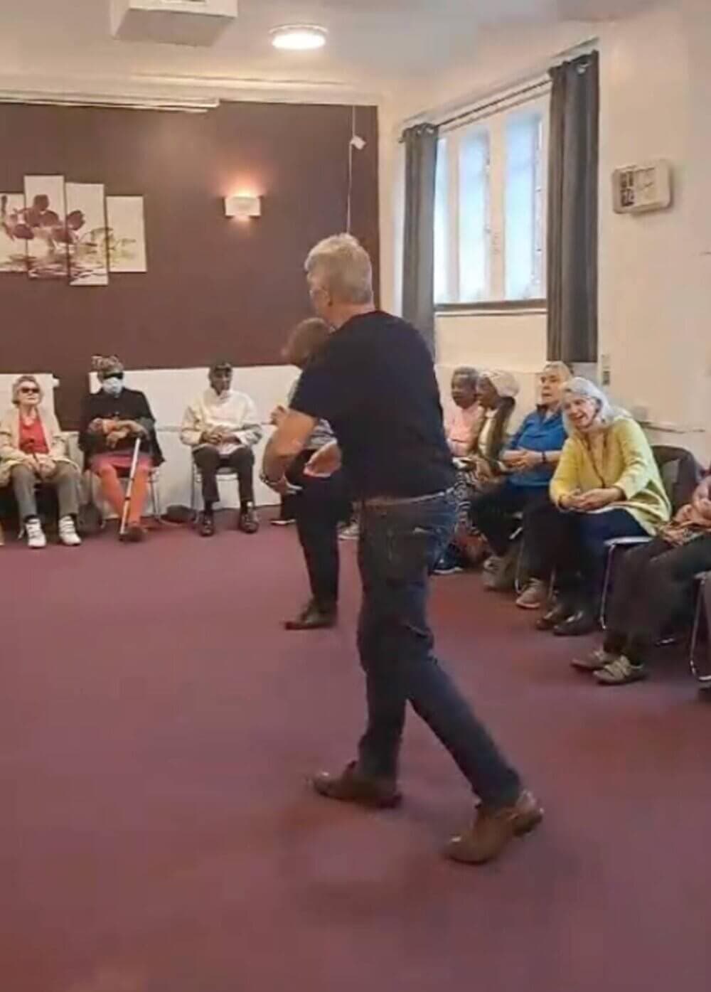 Two people dance in a room while others sit in a circle watching them. - Home Instead