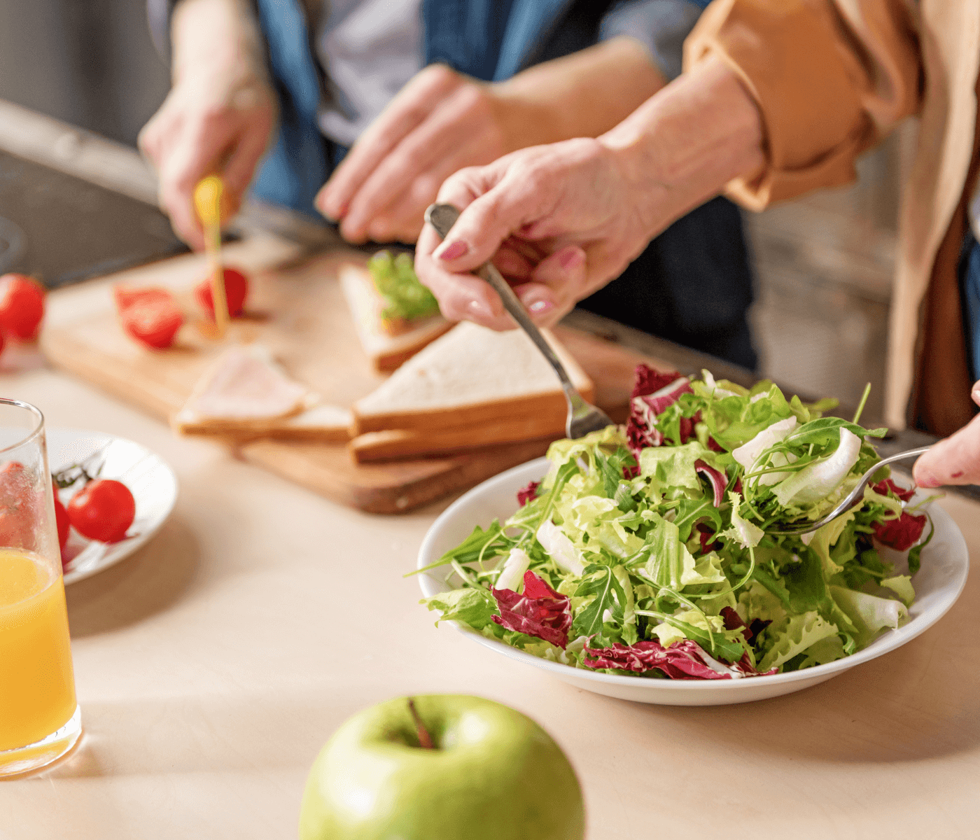 Two people preparing a meal; one making a salad and the other assembling a sandwich with orange juice and an apple nearby. - Home Instead