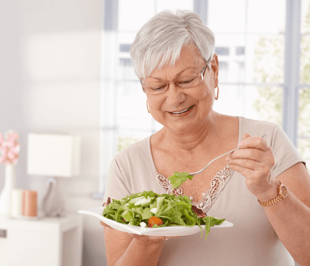 Elderly woman with short white hair smiling while holding a fork and eating a salad in a brightly lit room. - Home Instead