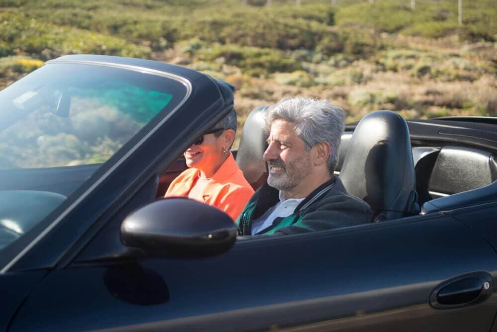 Two people smiling and driving in a black convertible car on a sunny day with green hills in the background. - Home Instead