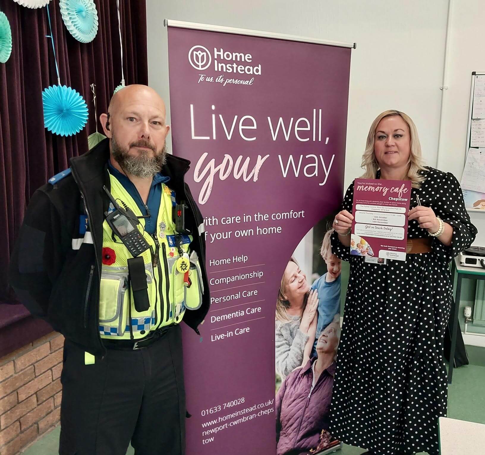 Two people stand beside a banner for Home Instead, promoting home care services. One holds a flyer for a memory café. - Home Instead