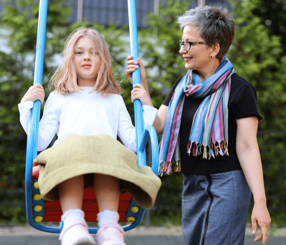 An older woman pushes a young girl on a swing in a park. The girl smiles while the woman looks at her. - Home Instead