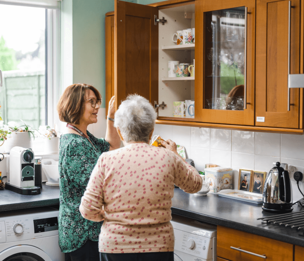 Two women, one elderly, engaging in conversation while standing in a kitchen. One is preparing items from a cabinet. - Home Instead