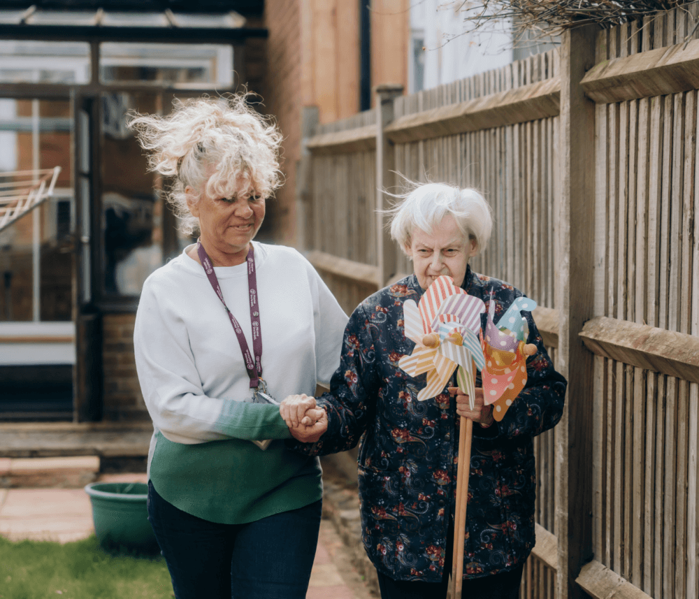 A caregiver and an elderly woman with a cane holding colorful pinwheels walk together in a garden. - Home Instead