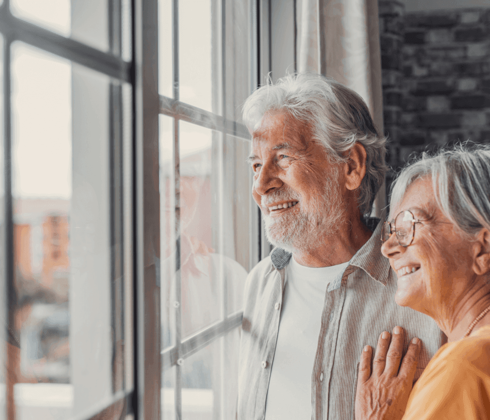 An elderly couple smiling and looking out of a window at something outside. The woman has her hand on the man's shoulder. - Home Instead