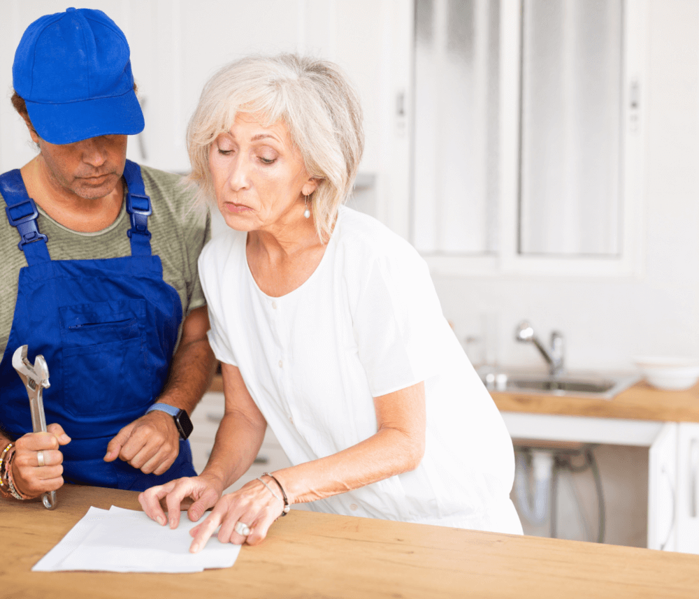 An older woman and a repairman review a document in a kitchen; the repairman holds a wrench. - Home Instead