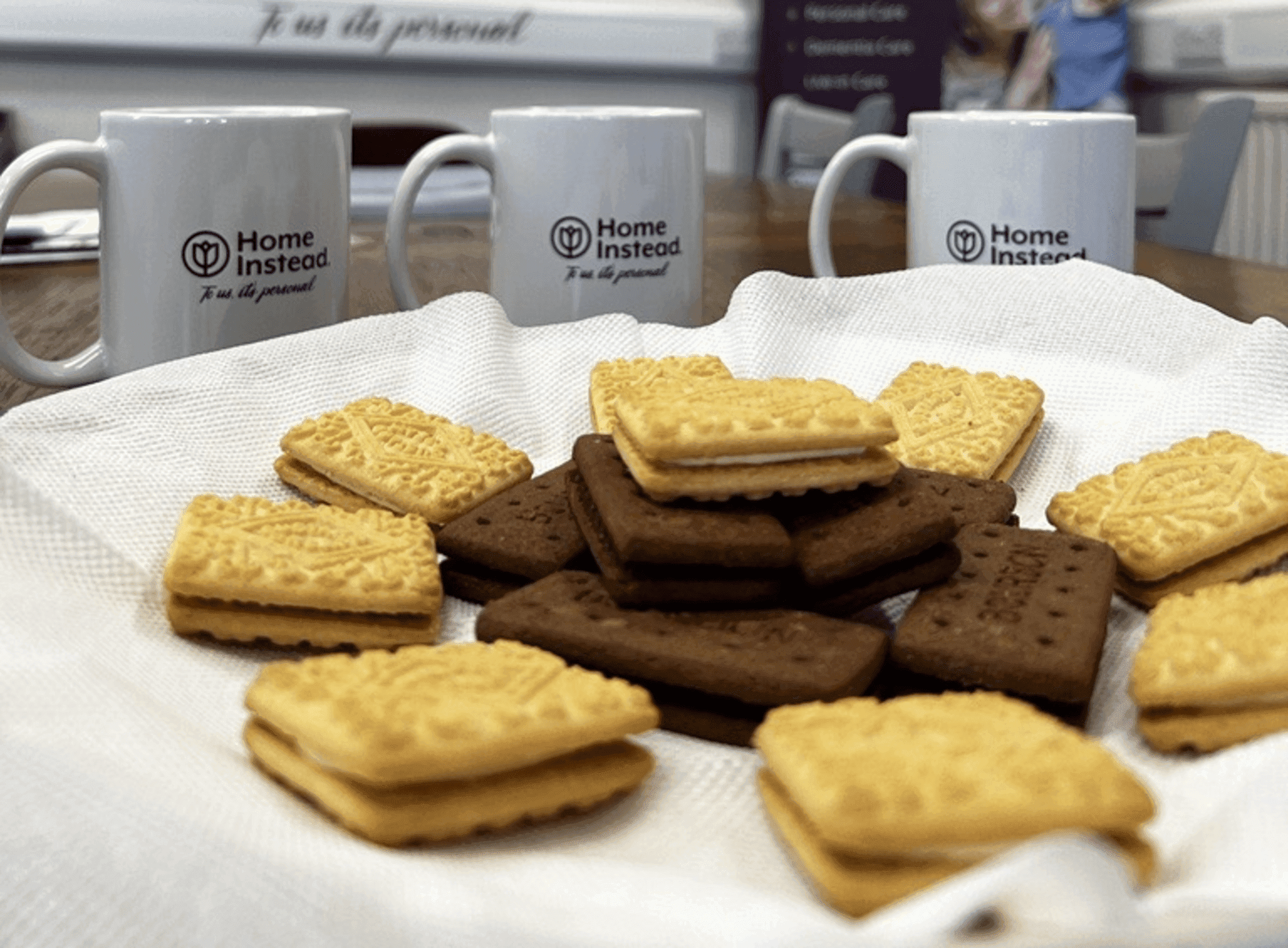 Assorted biscuits on a napkin with three "Home Instead" mugs in the background. - Home Instead