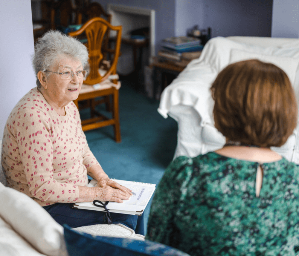 Two women sit in a living room, engaged in conversation. An older woman holds a paper on her lap. - Home Instead