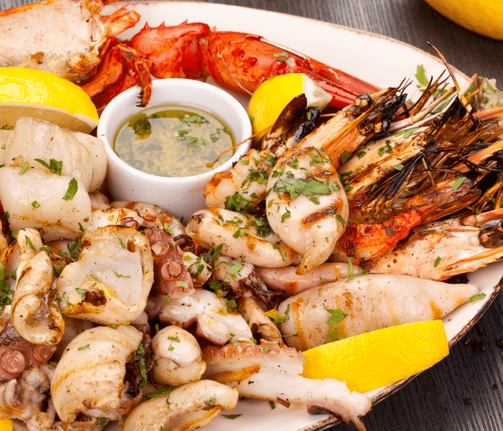 A seafood platter with grilled shrimp, octopus, squid, scallops, lobster, lemon wedges, and a dipping sauce. - Home Instead
