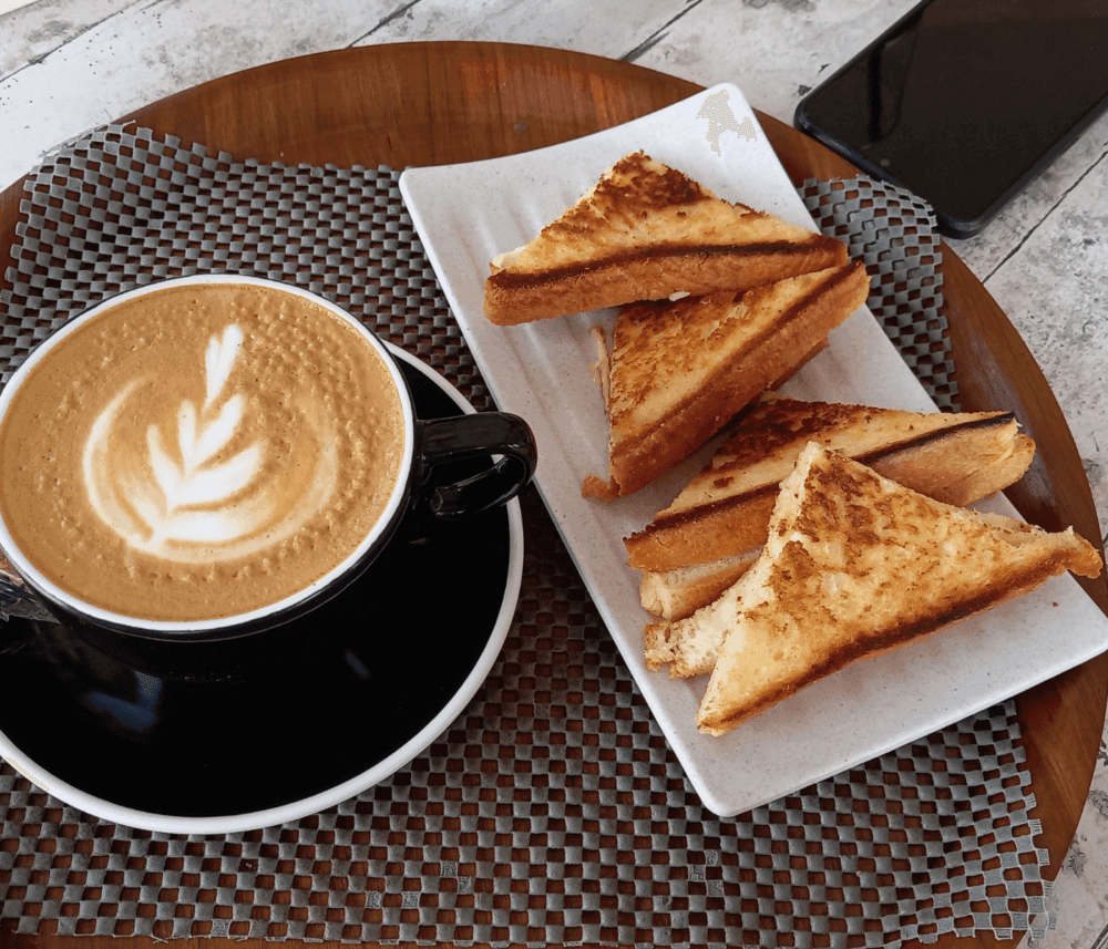 A latte with latte art in a black cup next to a plate of grilled cheese sandwich triangles on a wooden table. - Home Instead