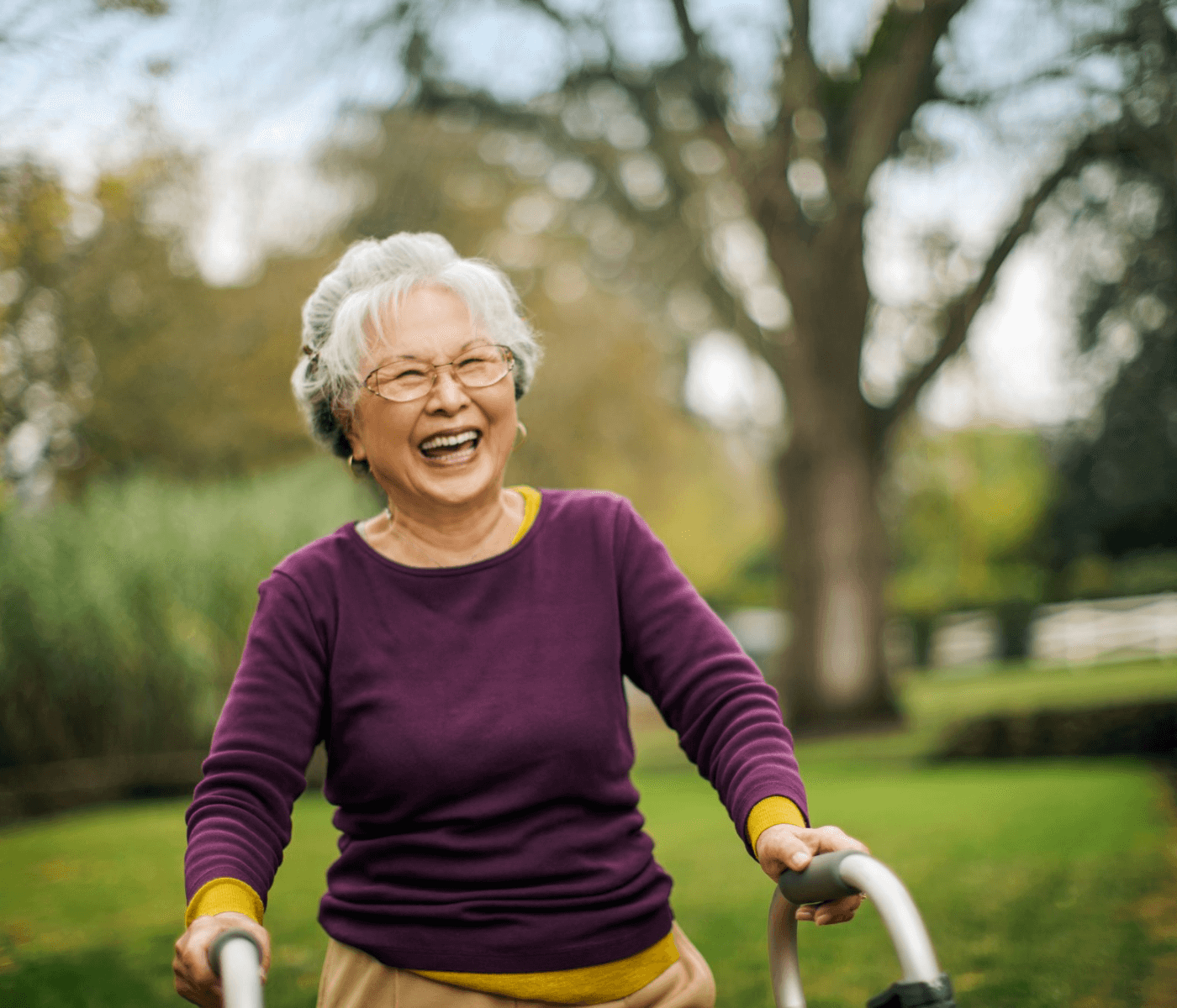 An elderly woman with a walker smiles joyfully in a park with trees in the background. - Home Instead
