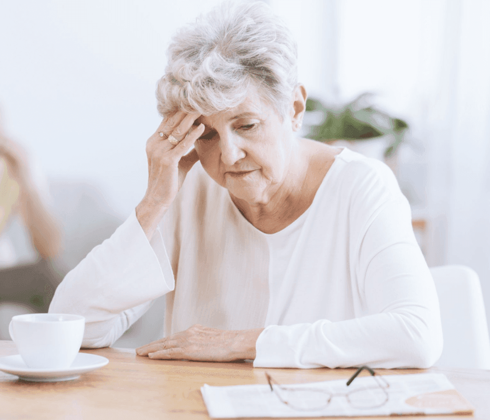 Elderly woman sitting at a table, holding her head in one hand with a concerned expression, a cup and paper in front of her. - Home Instead