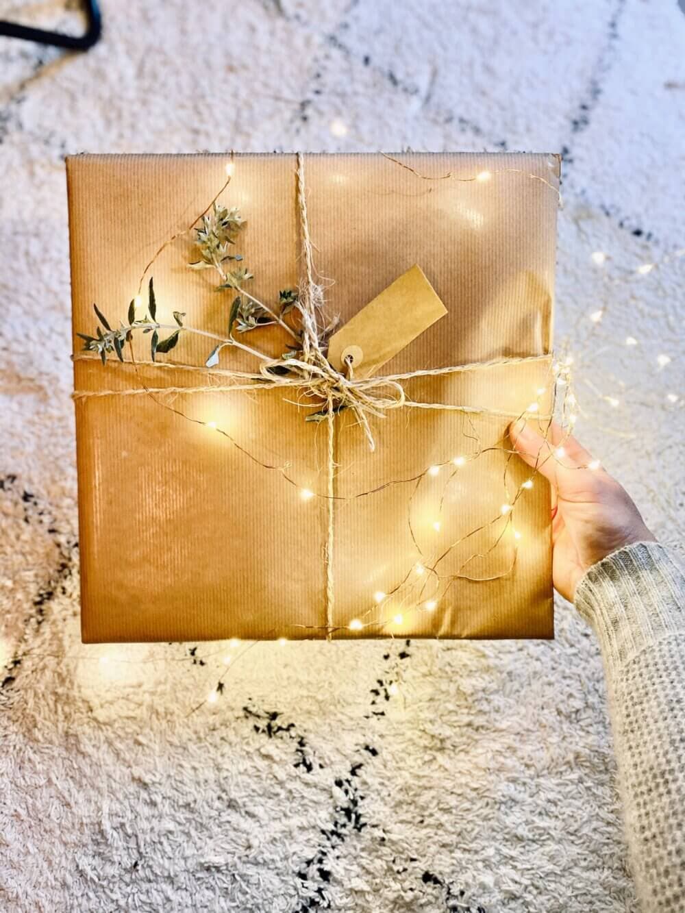 A hand holds a gift wrapped in brown paper, decorated with string lights, greenery, and string on a soft, textured surface. - Home Instead