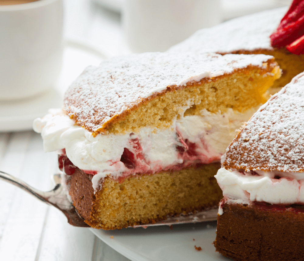 A close-up of a slice of Victoria sponge cake with cream and strawberry filling, dusted with powdered sugar. - Home Instead
