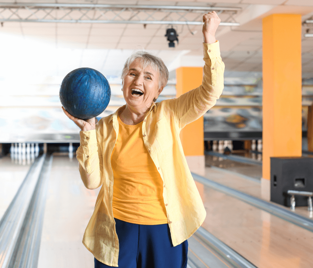 Elderly woman in a yellow shirt holding a blue bowling ball and cheering at a bowling alley. - Home Instead