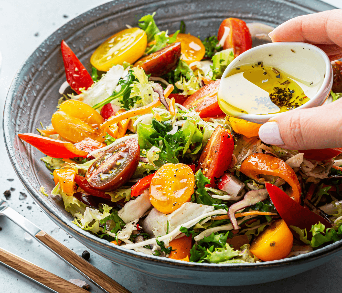 A bowl of fresh, colorful salad with tomatoes and greens being topped with olive oil and herbs from a small dish. - Home Instead