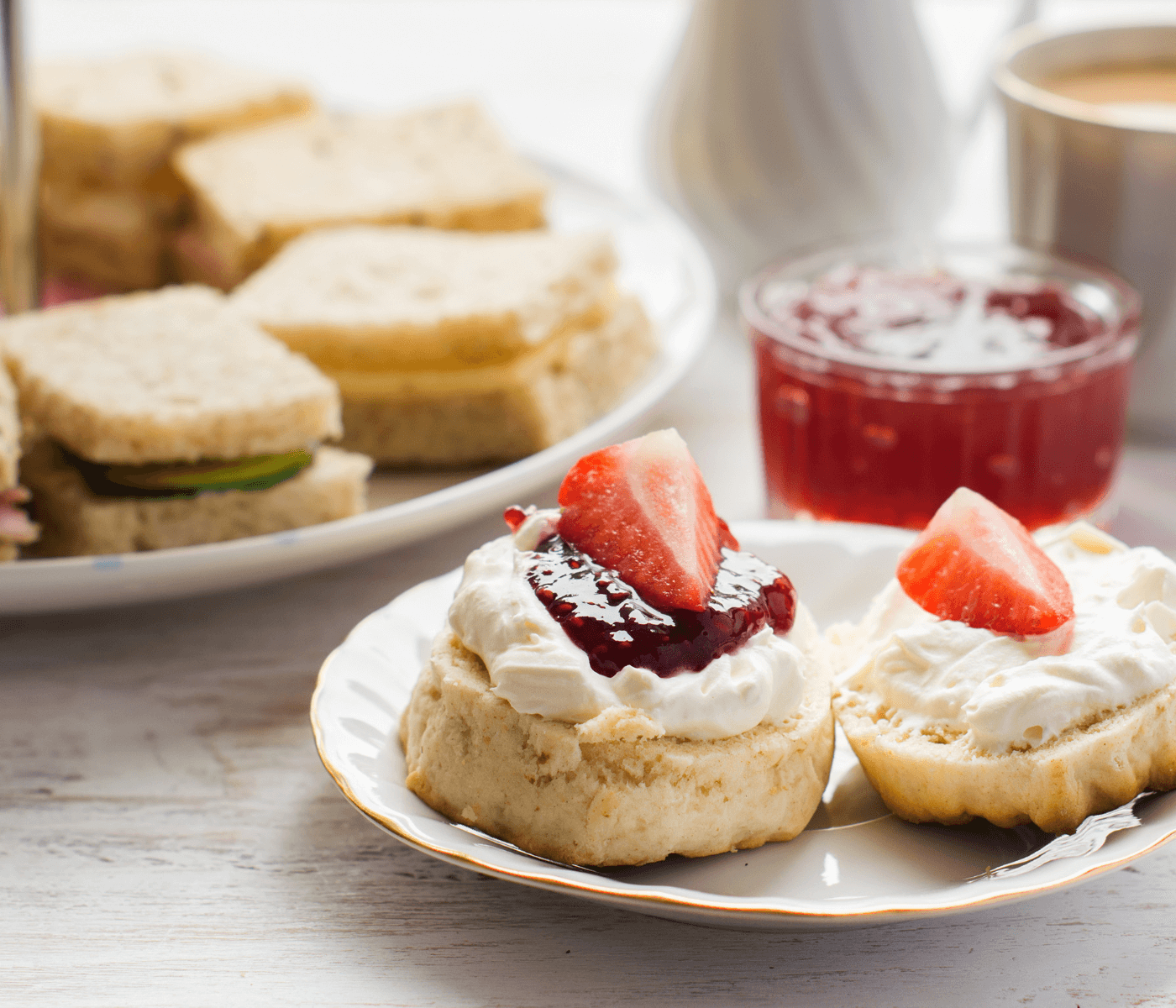 Scones with cream and strawberry jam, topped with fresh strawberries, served on a plate with a sandwich platter. - Home Instead