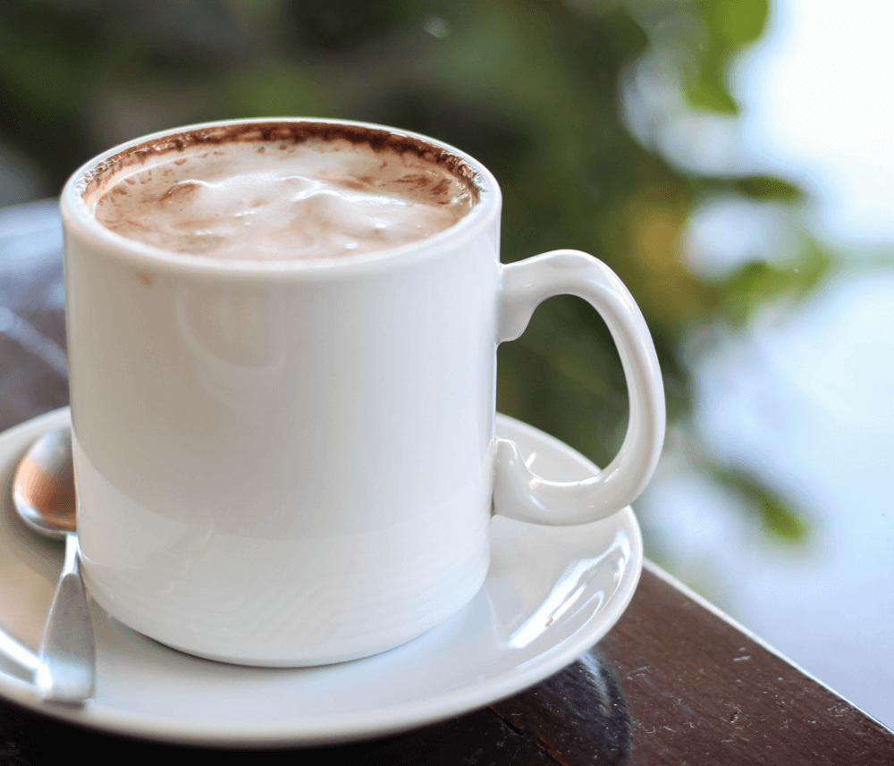 Close-up of a white coffee cup filled with a frothy beverage, placed on a saucer with a spoon, against a blurred background. - Home Instead