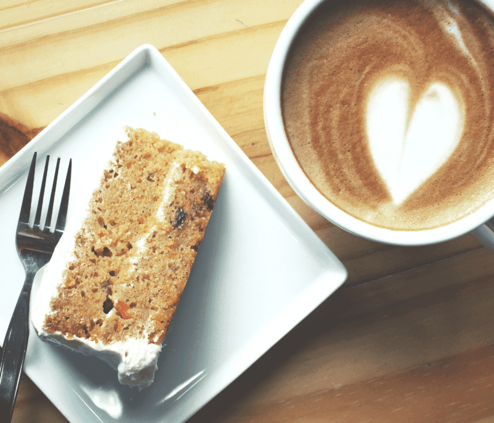 A slice of carrot cake next to a latte with a heart-shaped foam design on a wooden table. - Home Instead