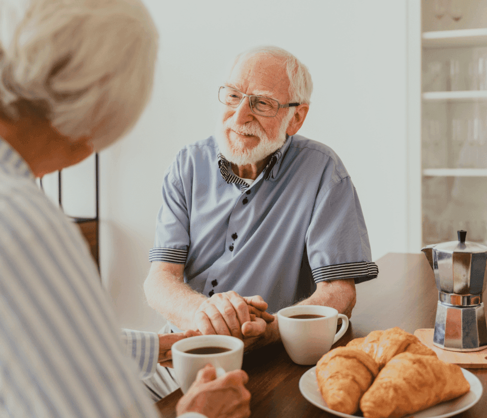 Elderly couple holding hands across a table with coffee and croissants, smiling and enjoying a moment together. - Home Instead