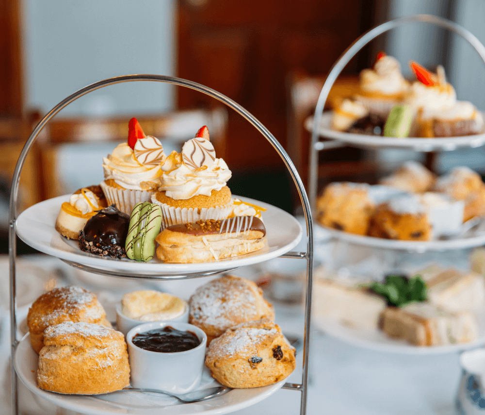 A tiered platter presents cupcakes, pastries, scones with cream and jam, and finger sandwiches for afternoon tea. - Home Instead