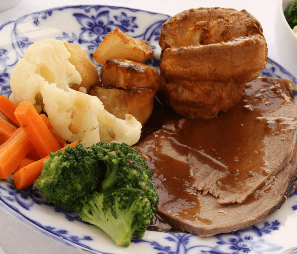 A plate with roast beef, Yorkshire pudding, potatoes, broccoli, carrots, and cauliflower, served with gravy. - Home Instead