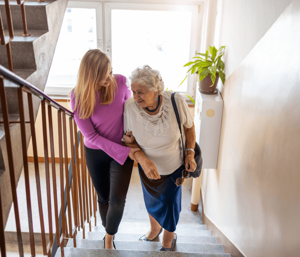 A young woman helps an elderly woman up the stairs in a bright stairwell, both smiling warmly. - Home Instead