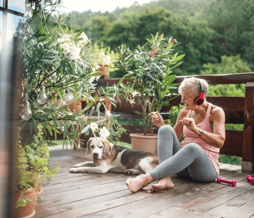 Senior woman with headphones sits on a wooden deck with her dog, surrounded by plants, enjoying music outdoors. - Home Instead