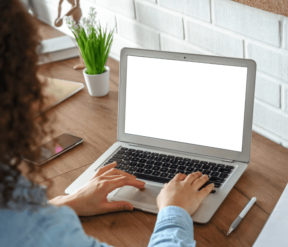 Person typing on a laptop with a blank white screen, seated at a wooden desk with a plant, phone, and pen nearby. - Home Instead