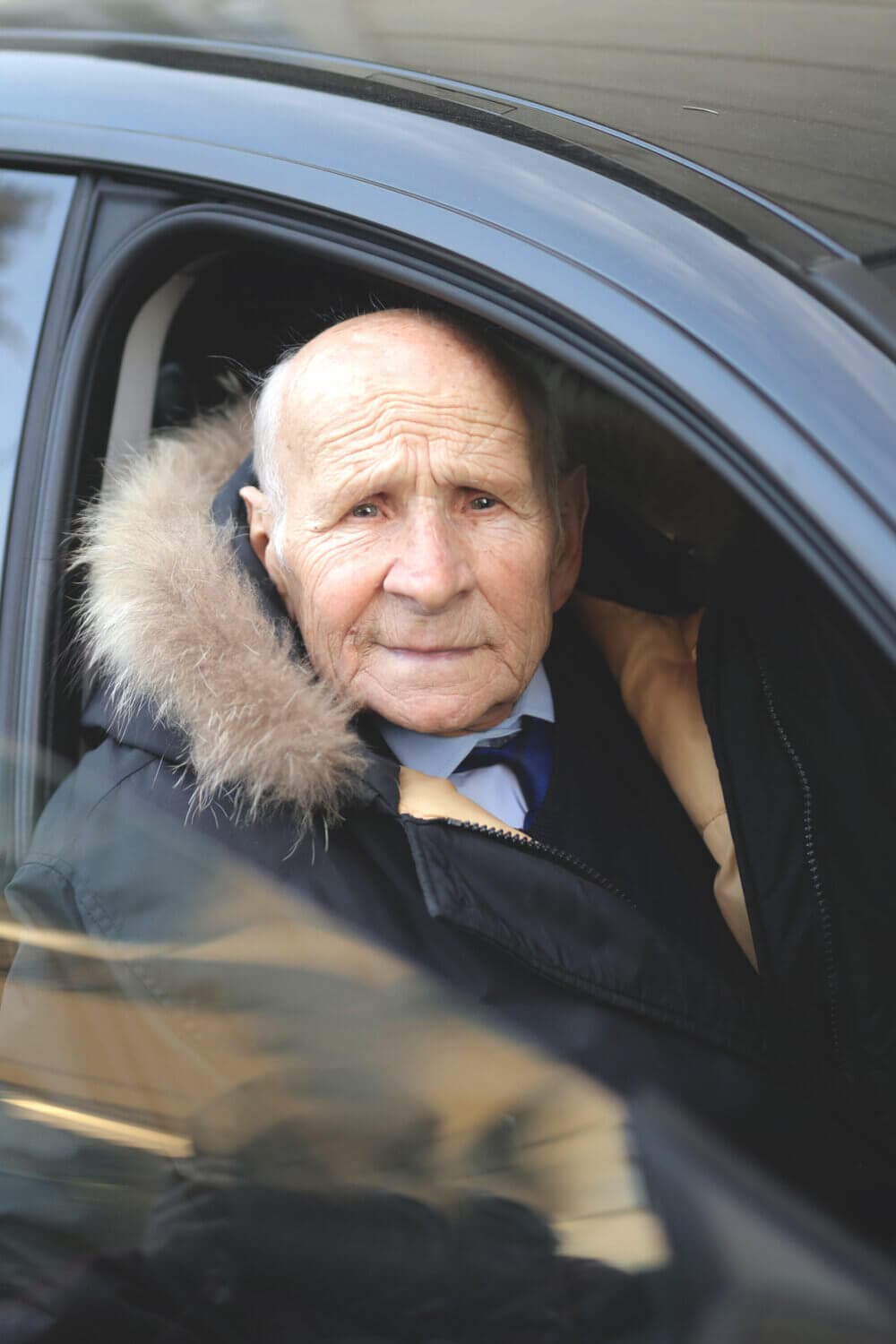 An elderly man wearing a fur-lined coat sits in the driver's seat of a car, looking directly at the camera. - Home Instead