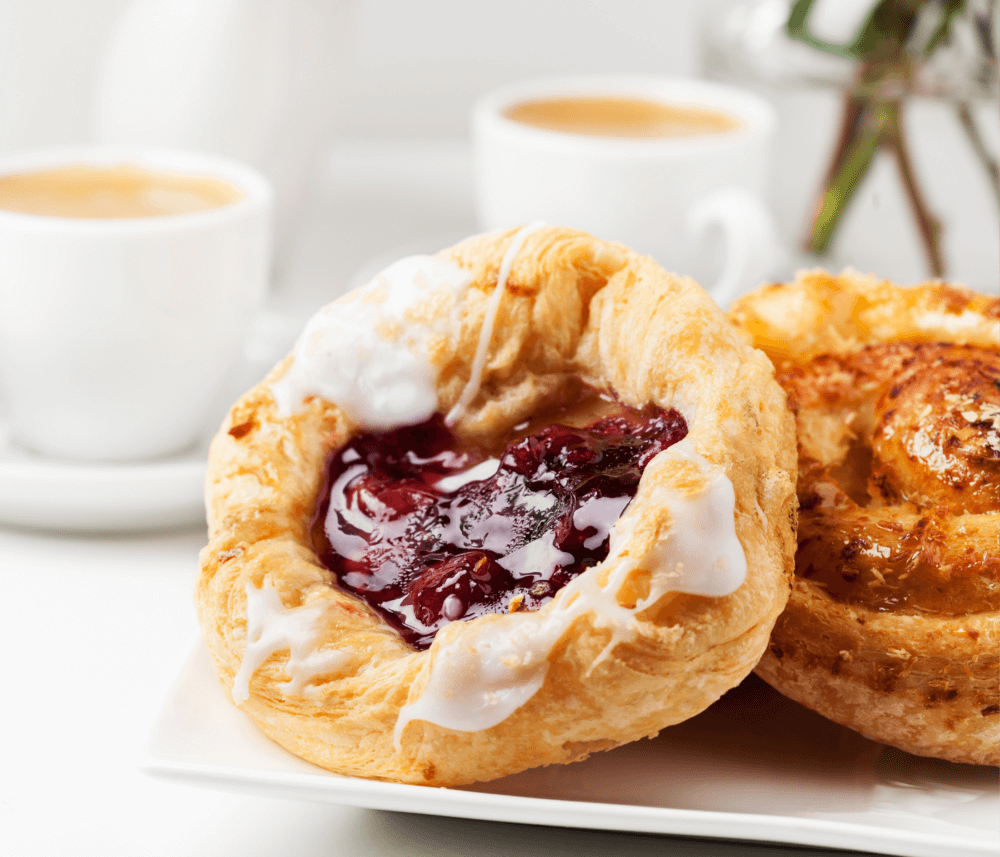 A plate with pastries, including one with a fruit filling and icing, accompanied by two cups of coffee in the background. - Home Instead