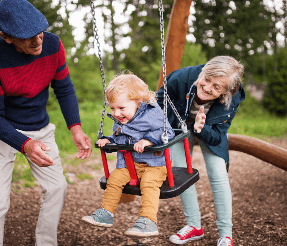 A child is joyfully swinging on a swing set, while two older adults happily push them at a park. - Home Instead