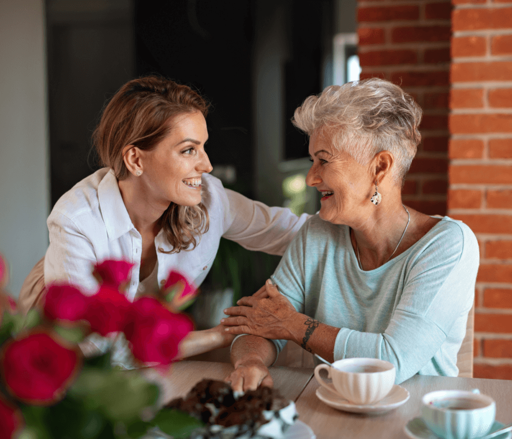 Two women sit at a table, smiling at each other, with a bouquet of flowers and coffee cups in the foreground. - Home Instead