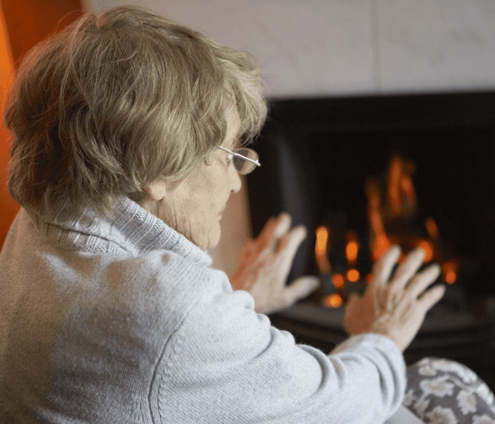 An elderly person with glasses sits by a fireplace, warming their hands in front of the flames. - Home Instead