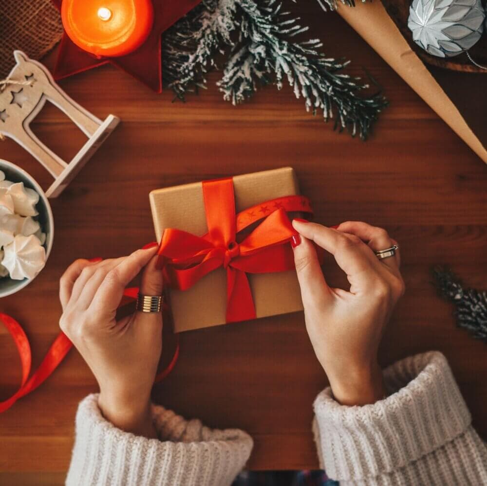Hands tying a red ribbon on a gift box, surrounded by festive decorations, candles, and pine branches. - Home Instead