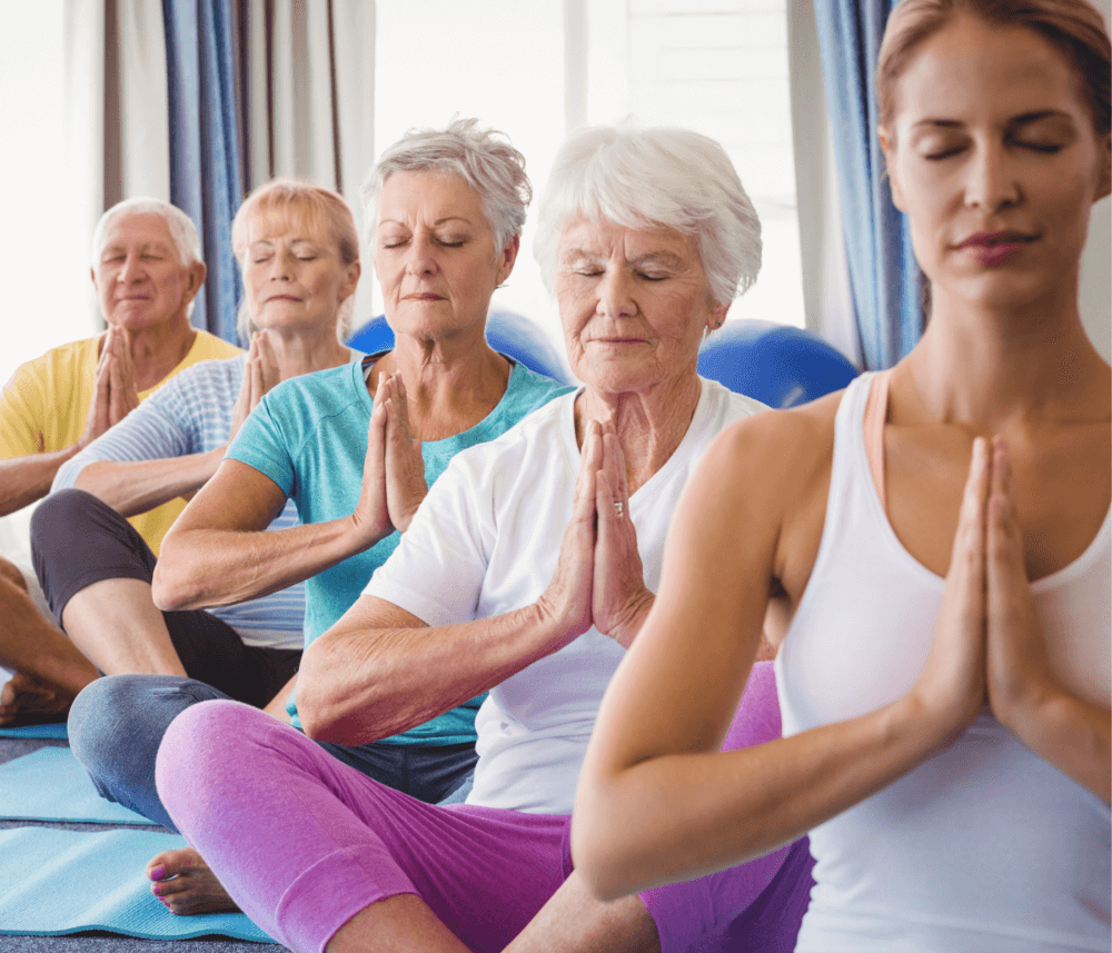 A group of elderly people and a younger woman meditate in a seated yoga pose with hands in prayer position. - Home Instead