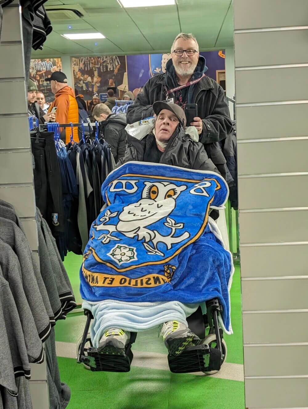 Two people in a store; one in a wheelchair with a blue and white blanket, the other standing behind and pushing the wheelchair. - Home Instead