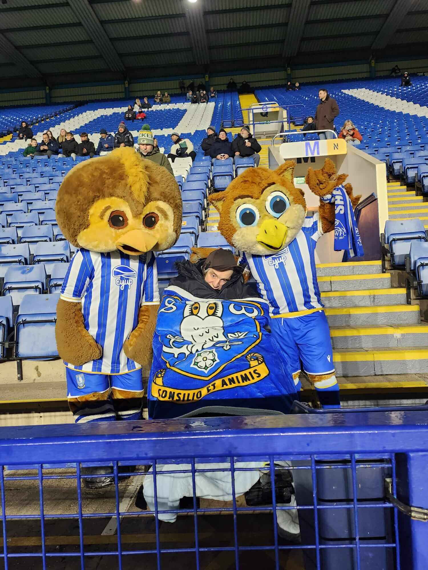 Two owl mascots in blue-and-white jerseys pose with a fan holding a club flag in a stadium. - Home Instead