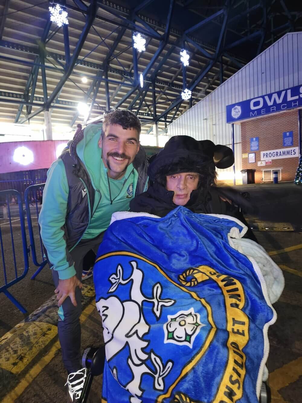 Two people pose for a photo at a stadium entrance, one is standing and the other in a wheelchair covered with a blanket. - Home Instead