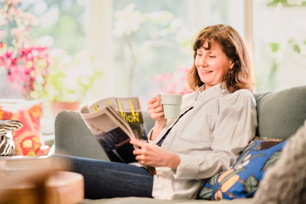 A woman sits on a couch, smiling while holding a cup of coffee and reading a magazine in a bright, cozy living room. - Home Instead