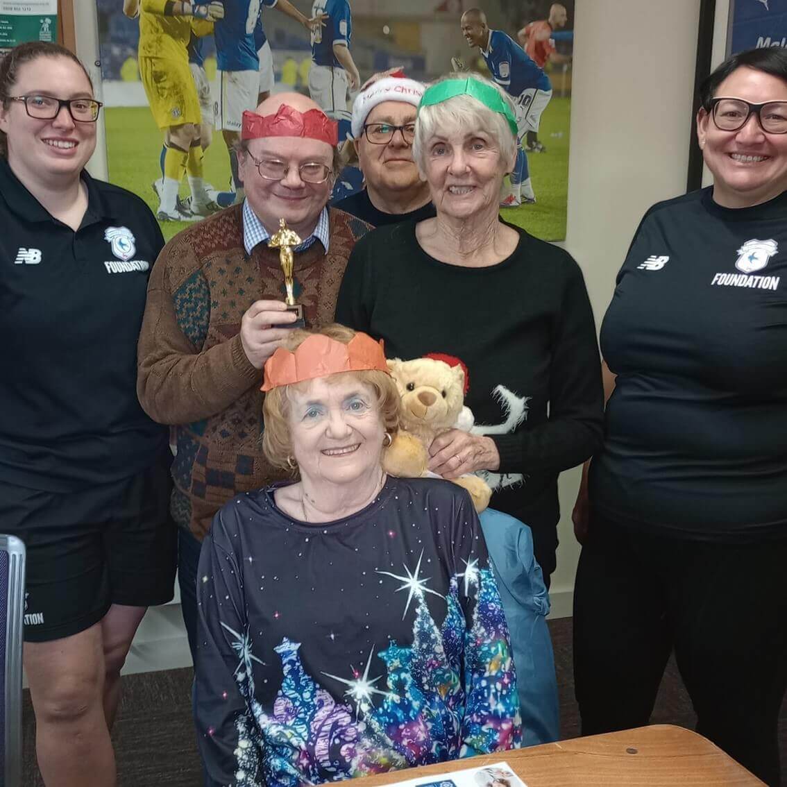 Group of six smiling people, some wearing festive hats, with one holding a trophy and another holding a teddy bear. - Home Instead