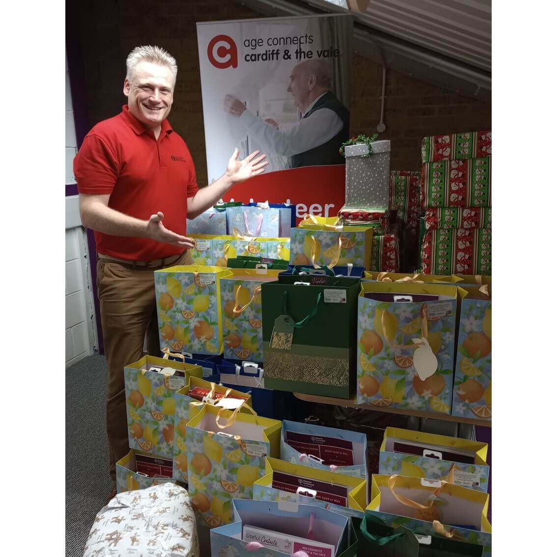 A man in a red shirt stands smiling beside a table filled with colorful gift bags and wrapped presents. - Home Instead