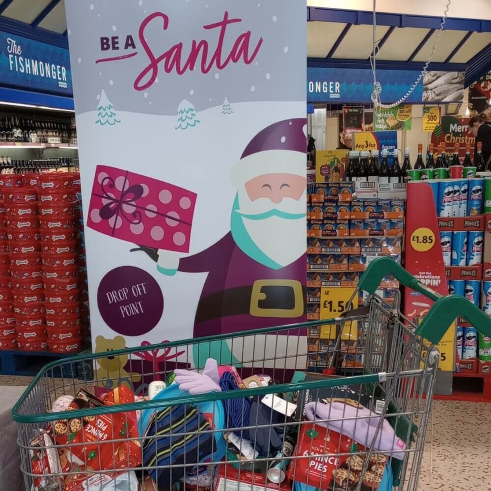 A shopping cart filled with donated items in front of a "Be a Santa" collection point at a grocery store. - Home Instead
