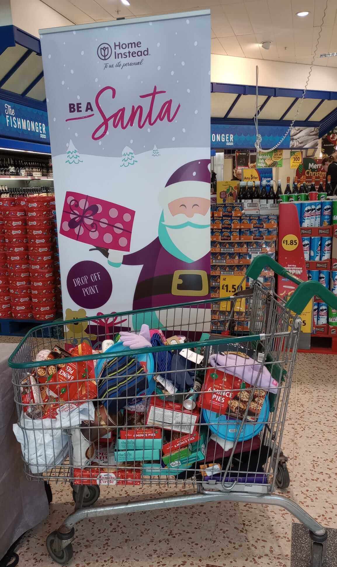 A shopping cart filled with gifts and items in front of a "Be a Santa" drop-off point sign at a supermarket. - Home Instead